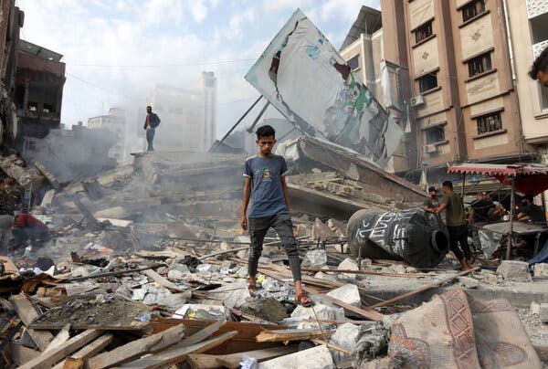 A youth stands amid rubble surrounded by damaged buildings, with thin smoke wafting in the air and a half-fallen billboard behind him.