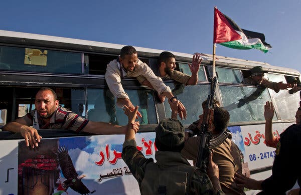 Former prisoners hang out the windows of a bus with Arabic writing while armed men outside grab their hands and wave a Palestinian flag.