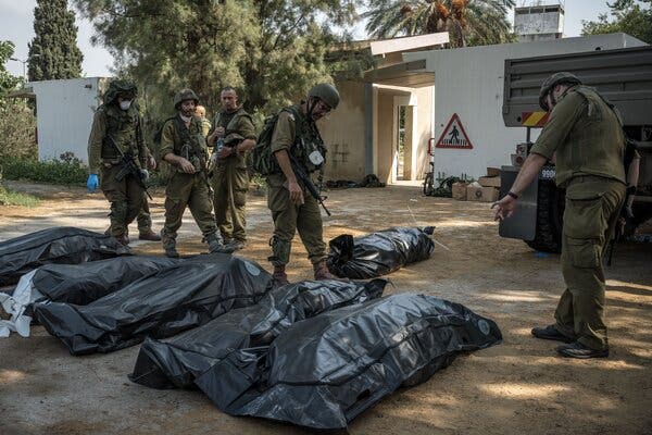 Armed soldiers stand around six black body bags lying on the ground near a one-story cream building with trees out front.