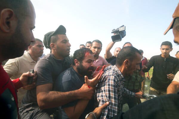 Men forcefully walking a bearded figure with blood on his forehead and hands as one person holds a video camera aloft.