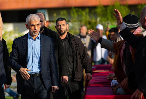 Yahya Sinwar, left forefront, in a blue shirt, suit and no tie, walks near a line of men raising their hands.
